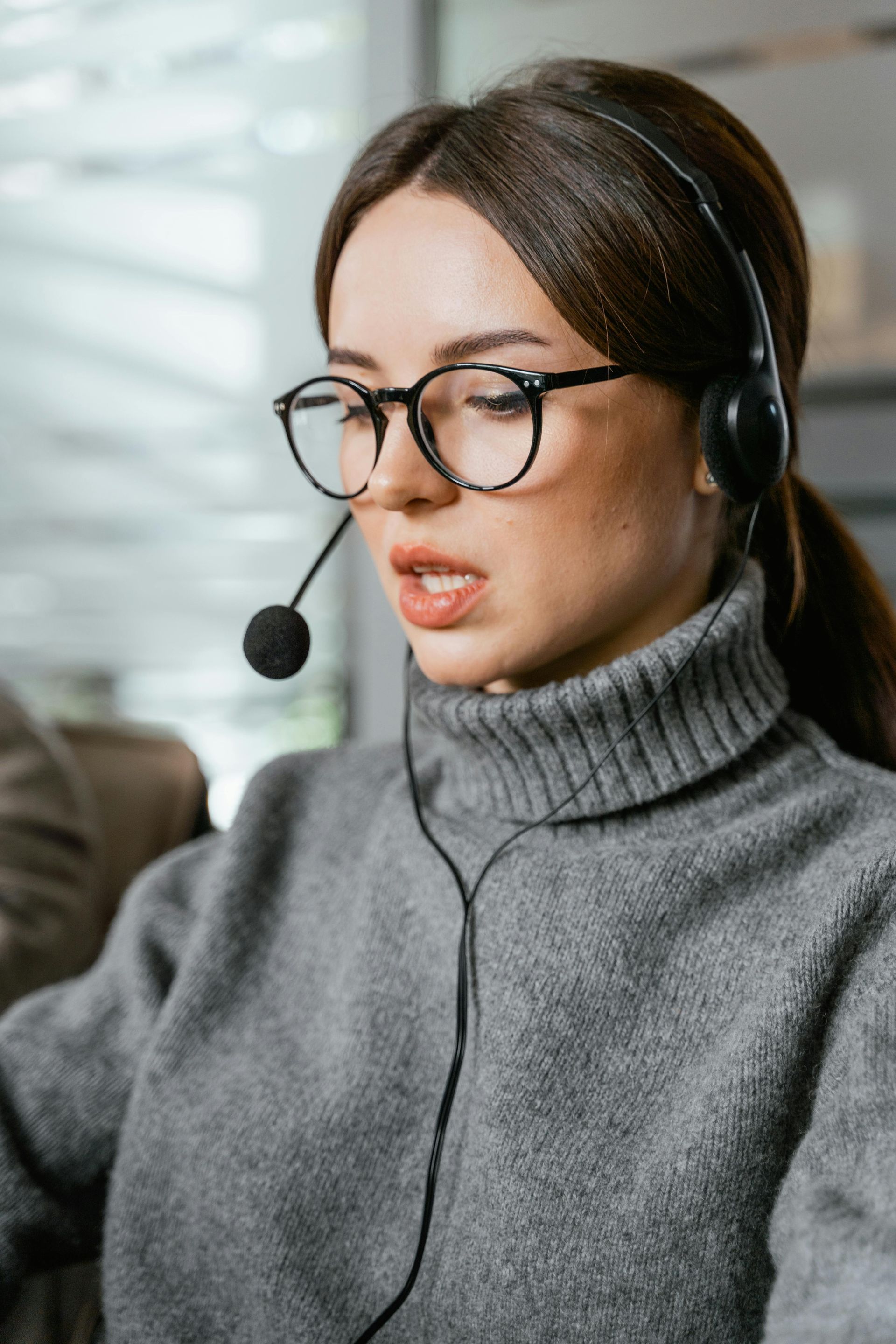 Woman wearing glasses and headset, speaking; gray turtleneck sweater.