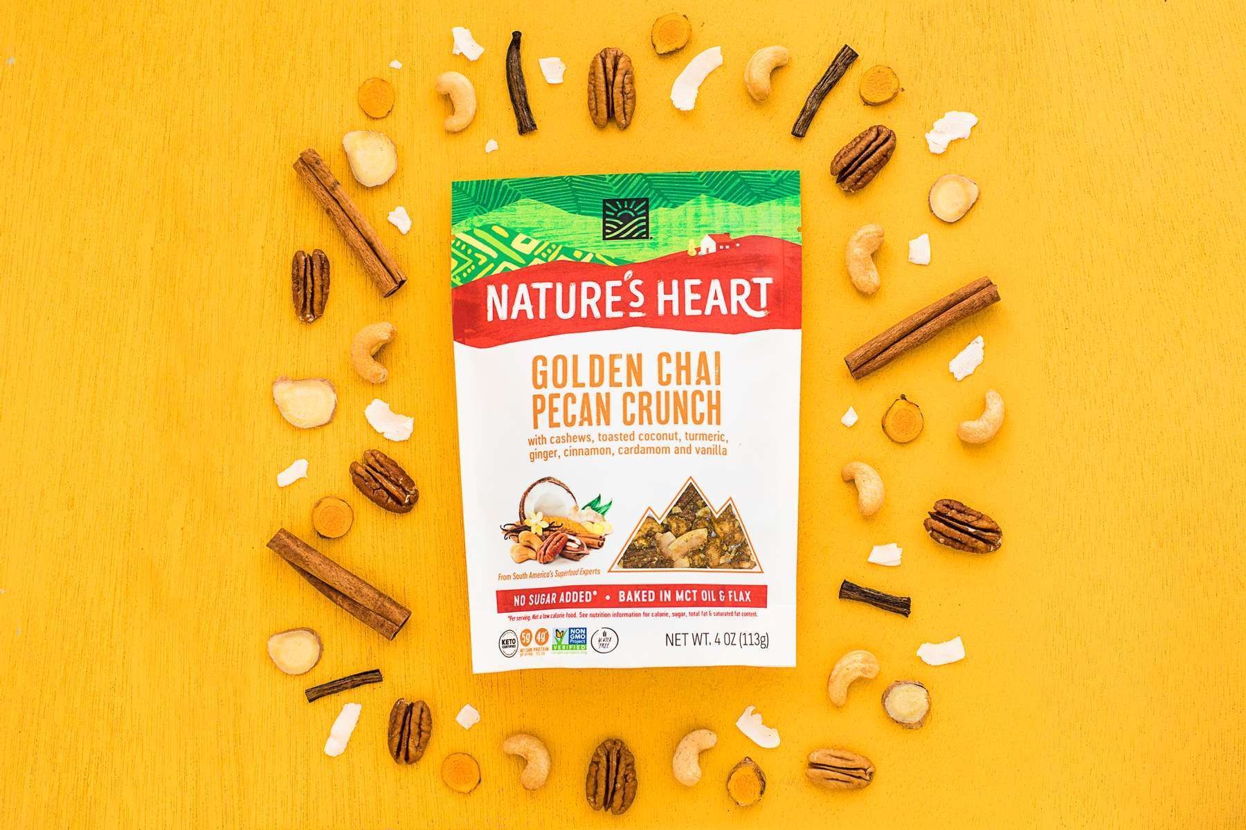 An assortment of walnuts, cashews, cinnamon sticks, coconut flakes and pecans on a golden yellow background, around Nature's Heart Golden Chai Pecan Crunch snack mix. Product photography for Nature's Heart by JAM Creative.