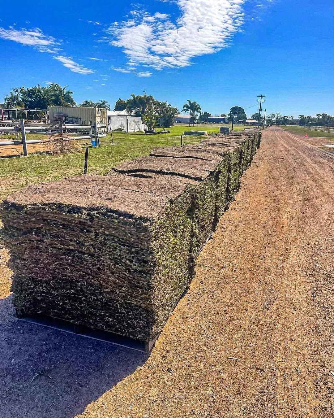 Stacks of Sod Lining a Dirt Road Under a Blue Sky, Grass Fields, and A Few Buildings — Top Notch Turf in Mackay, QLD