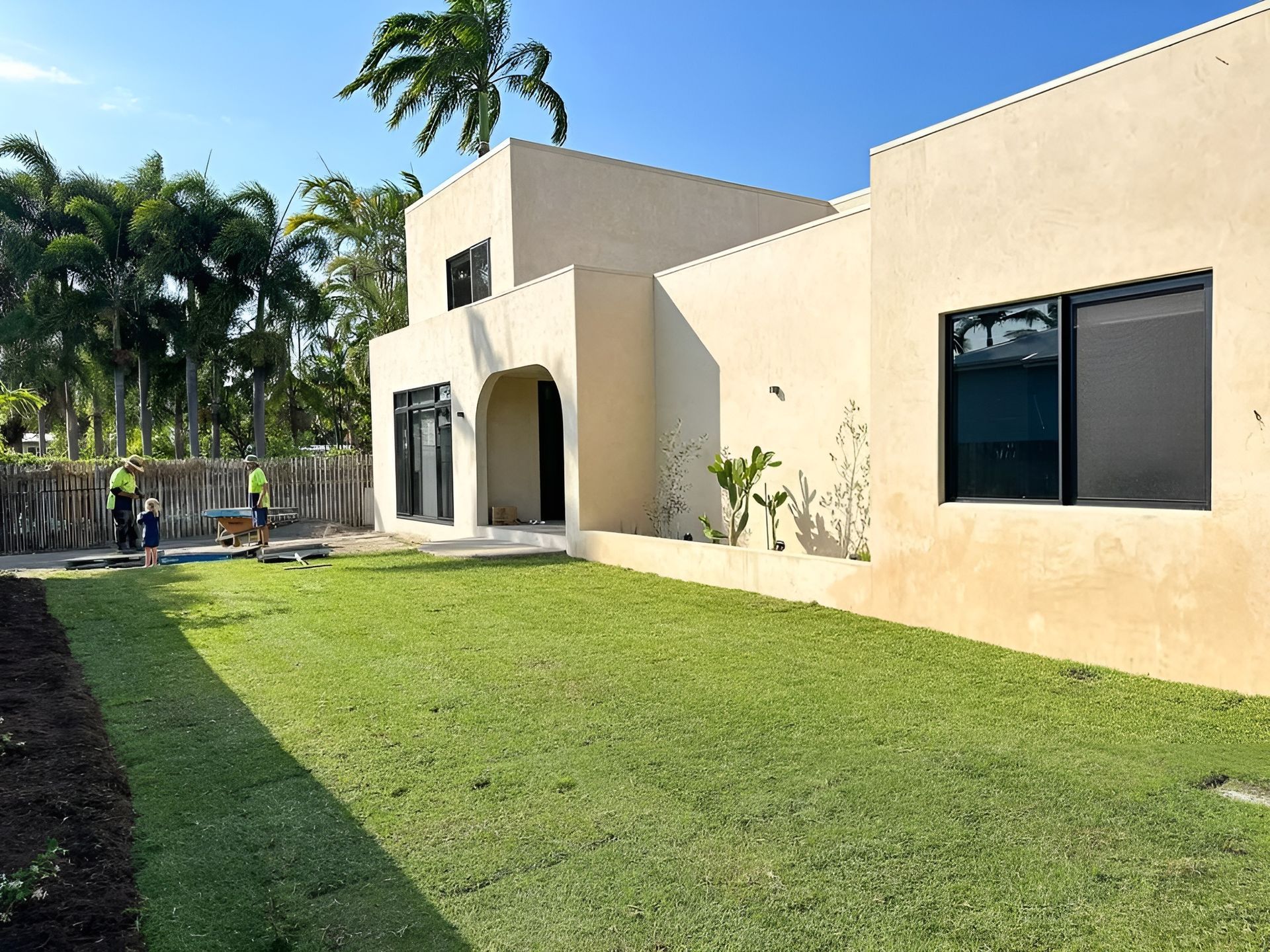 Beige Stucco House with Large Windows, Green Lawn, and Workers in The Distance — Top Notch Turf in Ayr, QLD