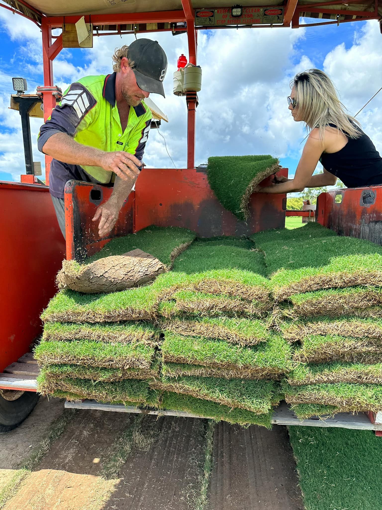 Two People Loading Squares of Grass Turf Into a Machine Under a Blue Sky — Top Notch Turf in Mount Isa, QLD