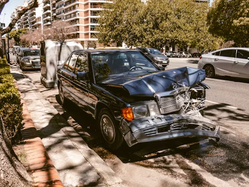 A dark sedan with significant front-end collision damage parked on a tree-lined city street.