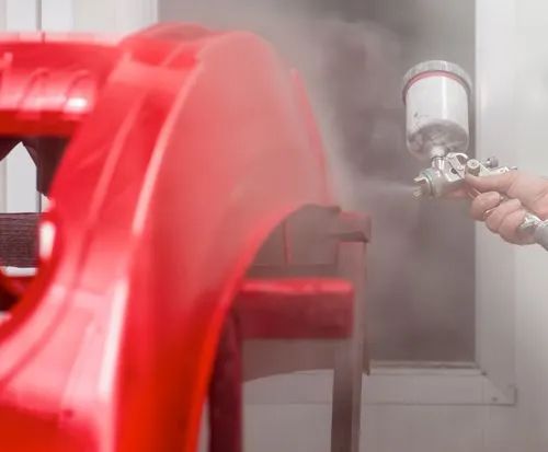 Spraying red painted metal part with a spray gun, creating a mist in a workshop.