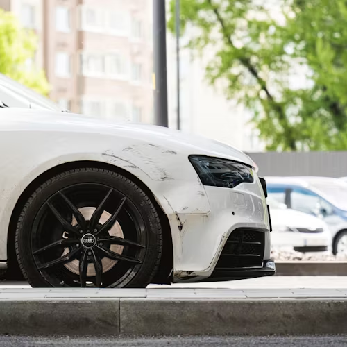 White car with black wheels and damage on front quarter panel, parked outdoors.