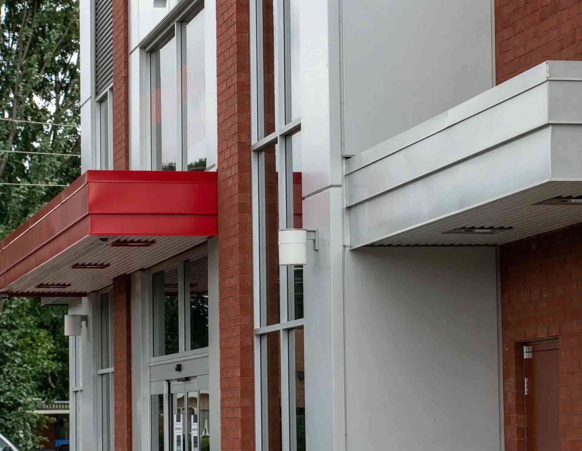 Une façade d'immeuble moderne caractérisée par un auvent d'entrée rouge, un avant-toit argenté, des colonnes en briques rouges et de grandes baies vitrées.