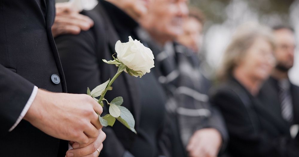 Person holding a white rose at a funeral; group of people in black attire in the background.