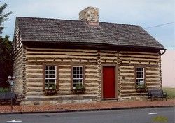 Log cabin with wooden shingle roof, three windows, red door, and stone chimney. Located on a brick lined street.