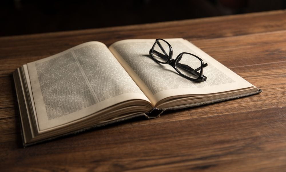 Open book with glasses resting on its pages, on a dark wooden surface.