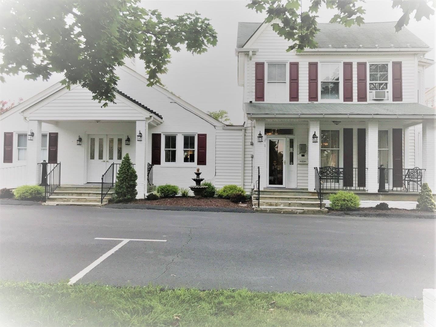 Two-story white building with red shutters.  Black asphalt parking lot in front, green grass and trees in the background.
