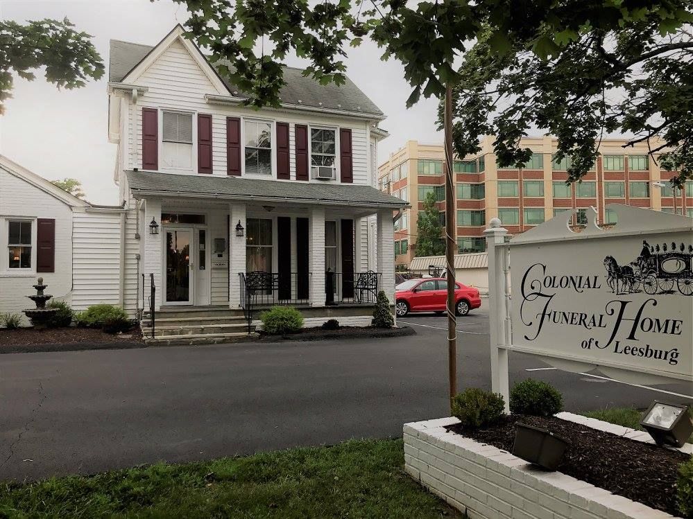 White colonial-style funeral home with red car parked out front. Sign on right: Colonial Funeral Home.