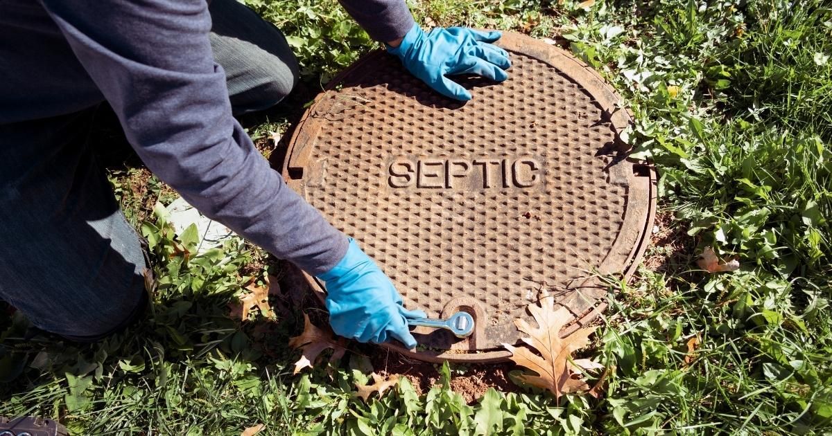Person in blue gloves opens a rusted septic tank cover in a grassy area.