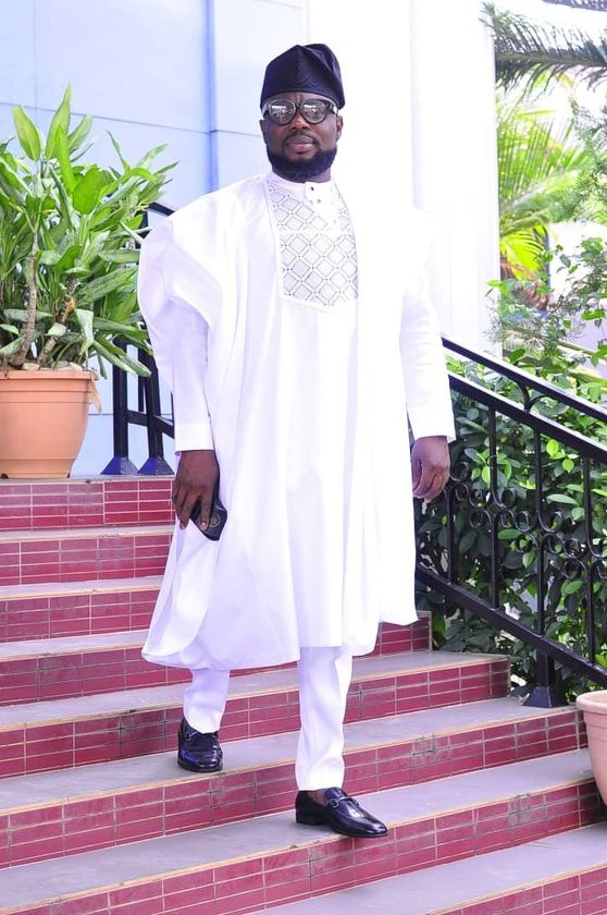 African man stood on red steps wearing a white agbada and black  oloye cap.