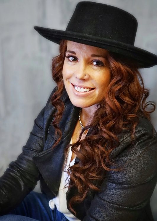 A woman with long red hair is wearing a black hat and smiling.