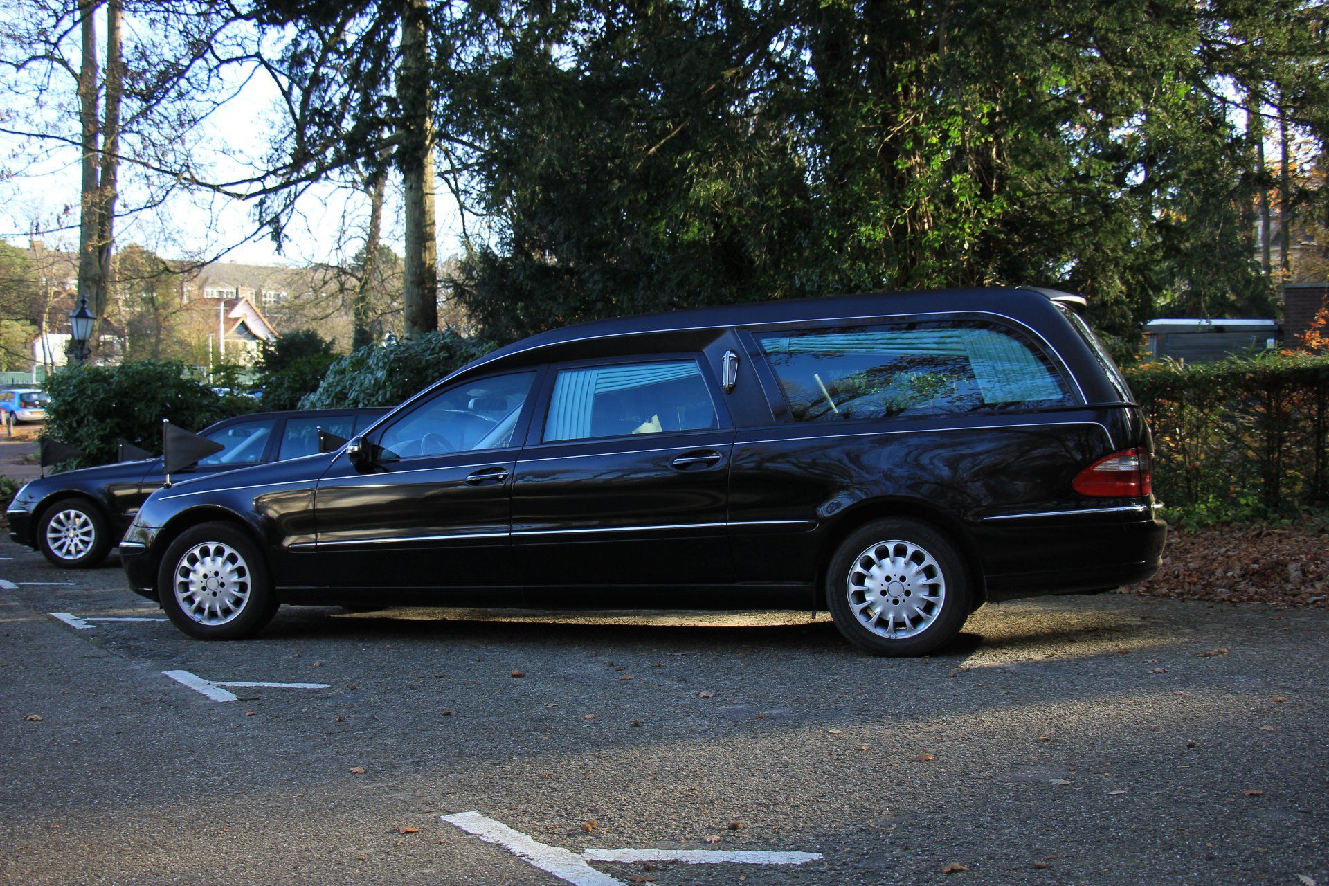 Una station wagon nera in stile carro funebre parcheggiata su una strada alberata accanto a un muro di pietra.