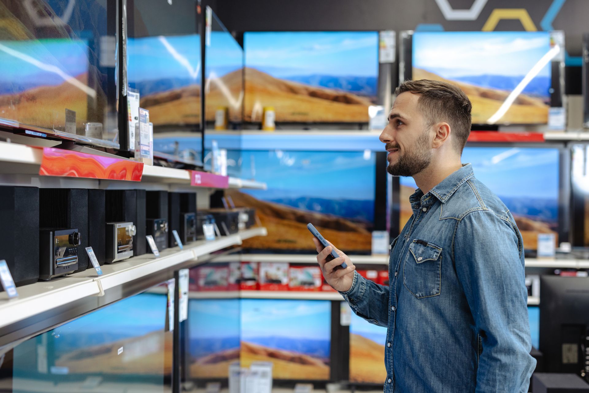 A man is looking at a television in a store while holding a cell phone.