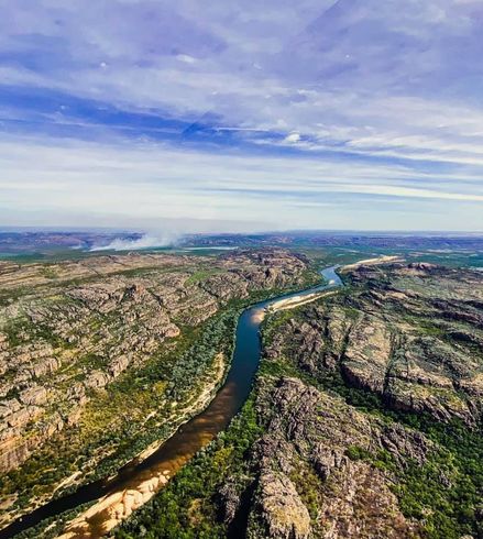 Aerial view of a winding river cutting through rocky green hills under a cloudy blue sky