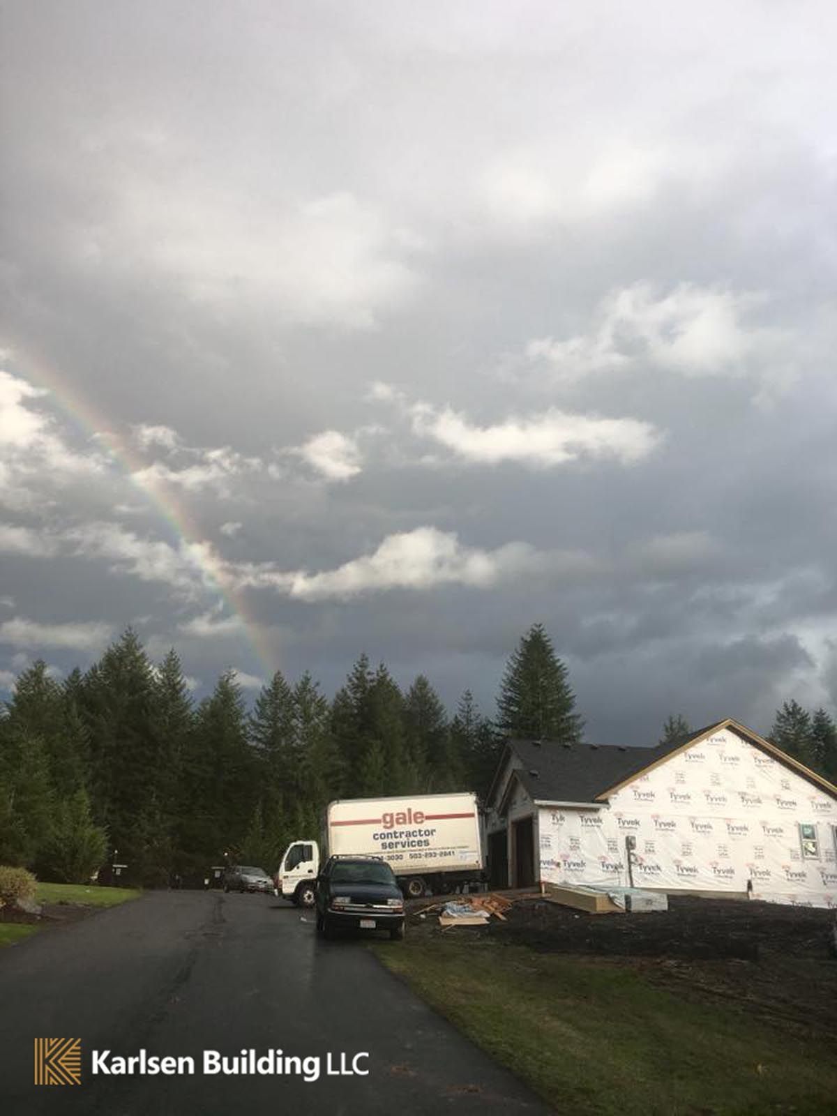 A truck is driving down a road next to a house with a rainbow in the sky.