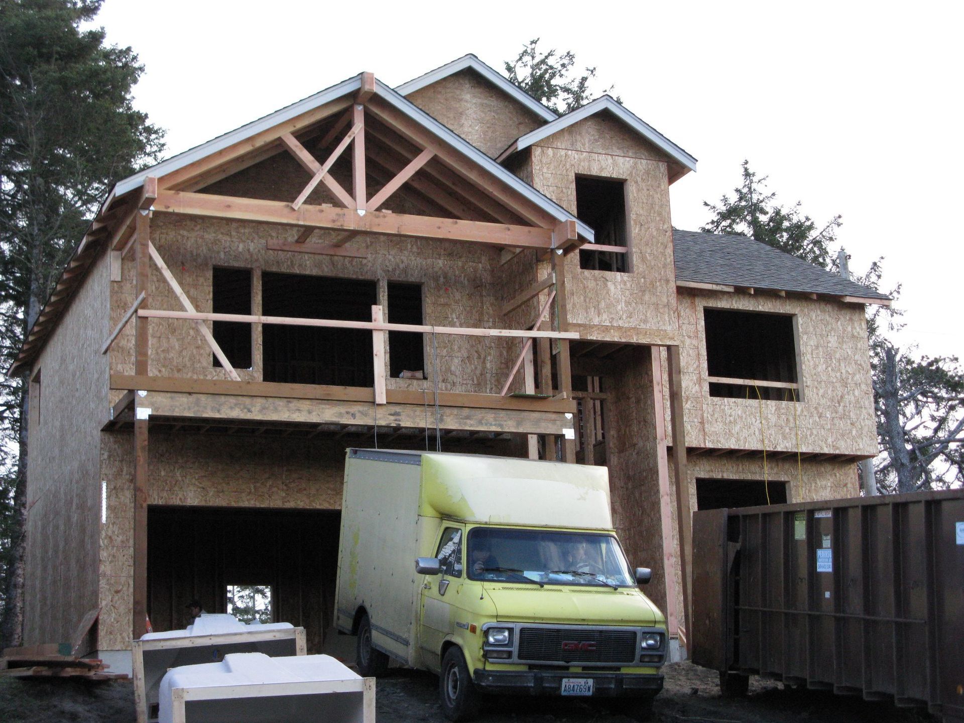 House under construction, unfinished wood structure. Yellow van parked in front, dumpster beside it.