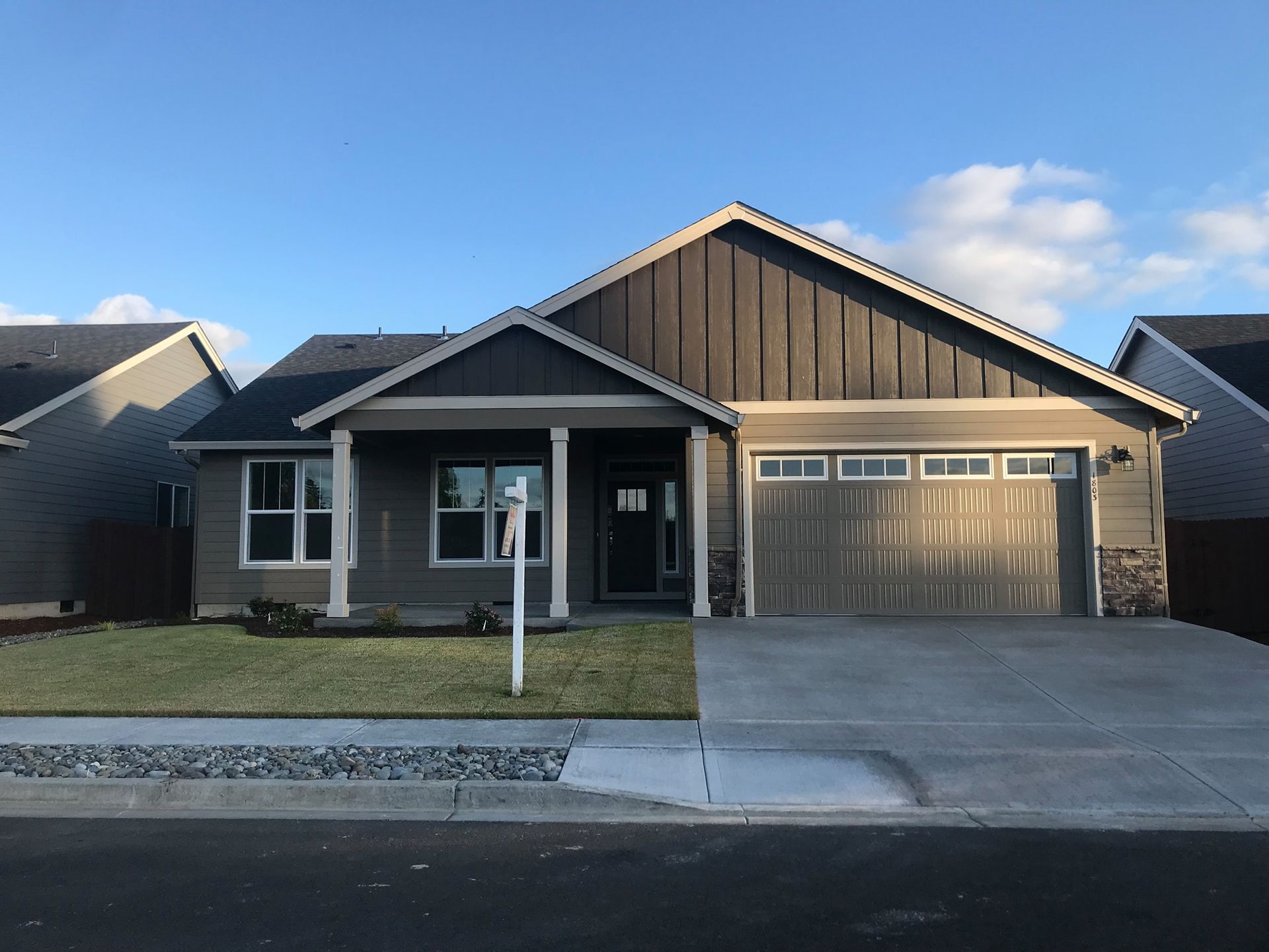 Gray ranch-style house with garage, grassy lawn, and sign in front of blue sky.