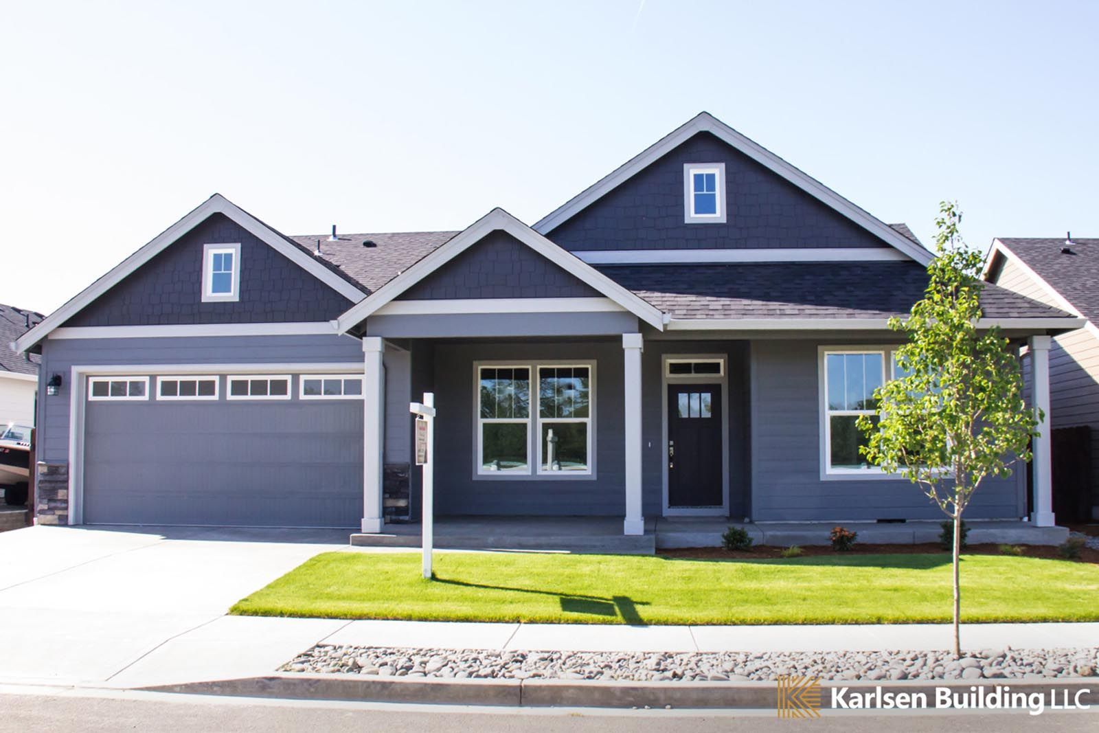 Blue house with gabled roof, garage, and front porch. Green lawn and a small tree in front.