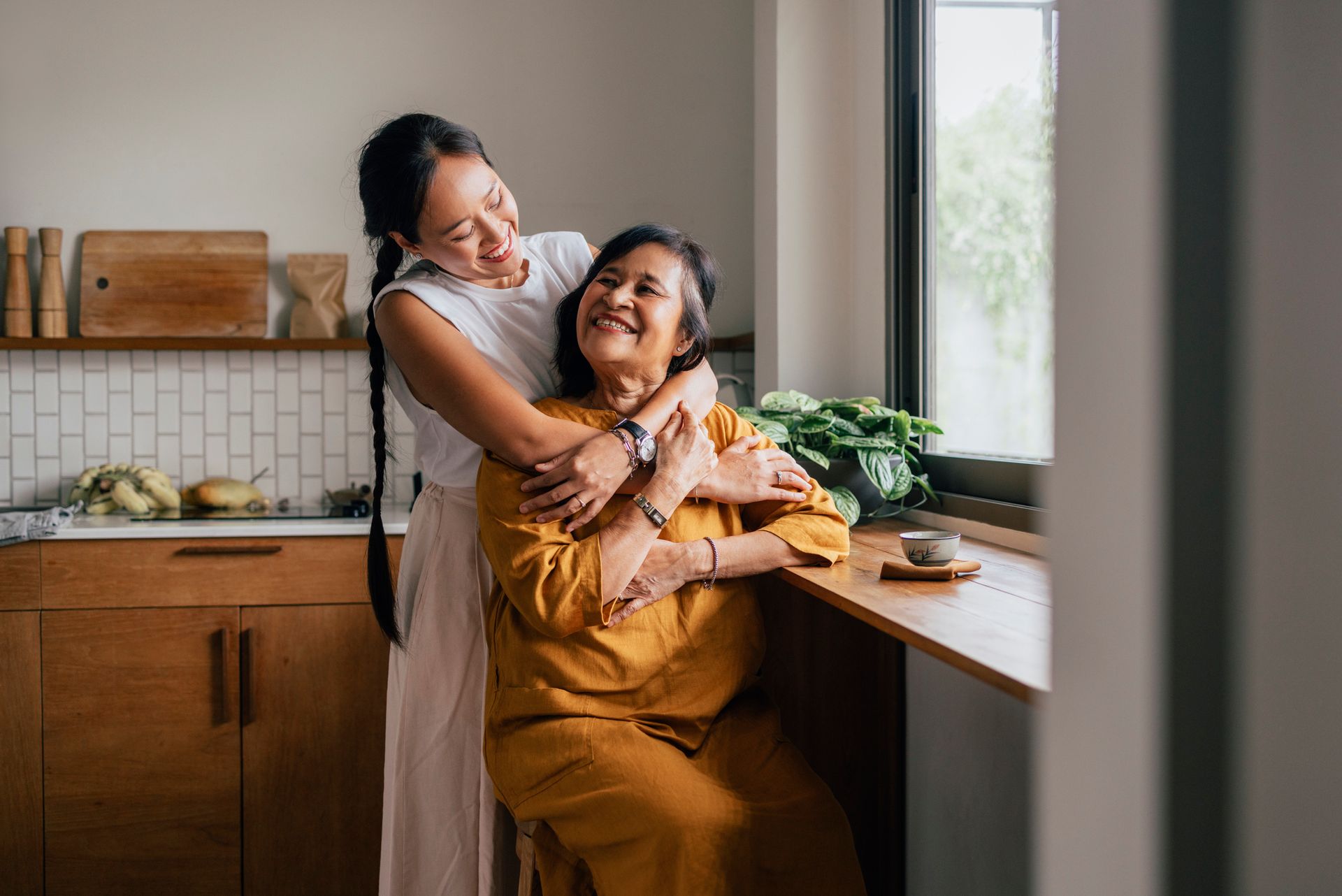 Woman embracing an older woman; both smiling in a kitchen by a window.