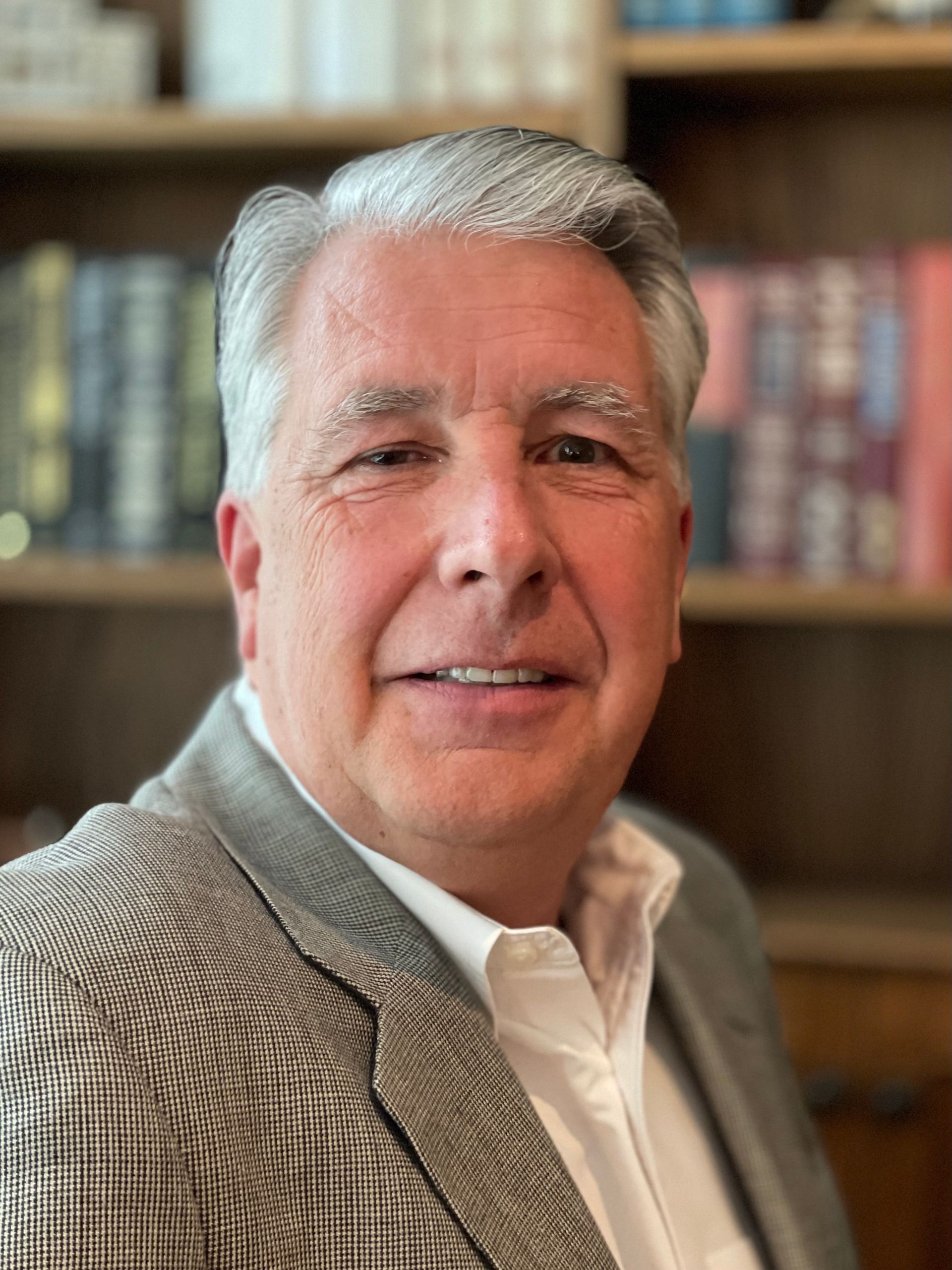 A man in a suit and white shirt is smiling in front of a bookshelf.