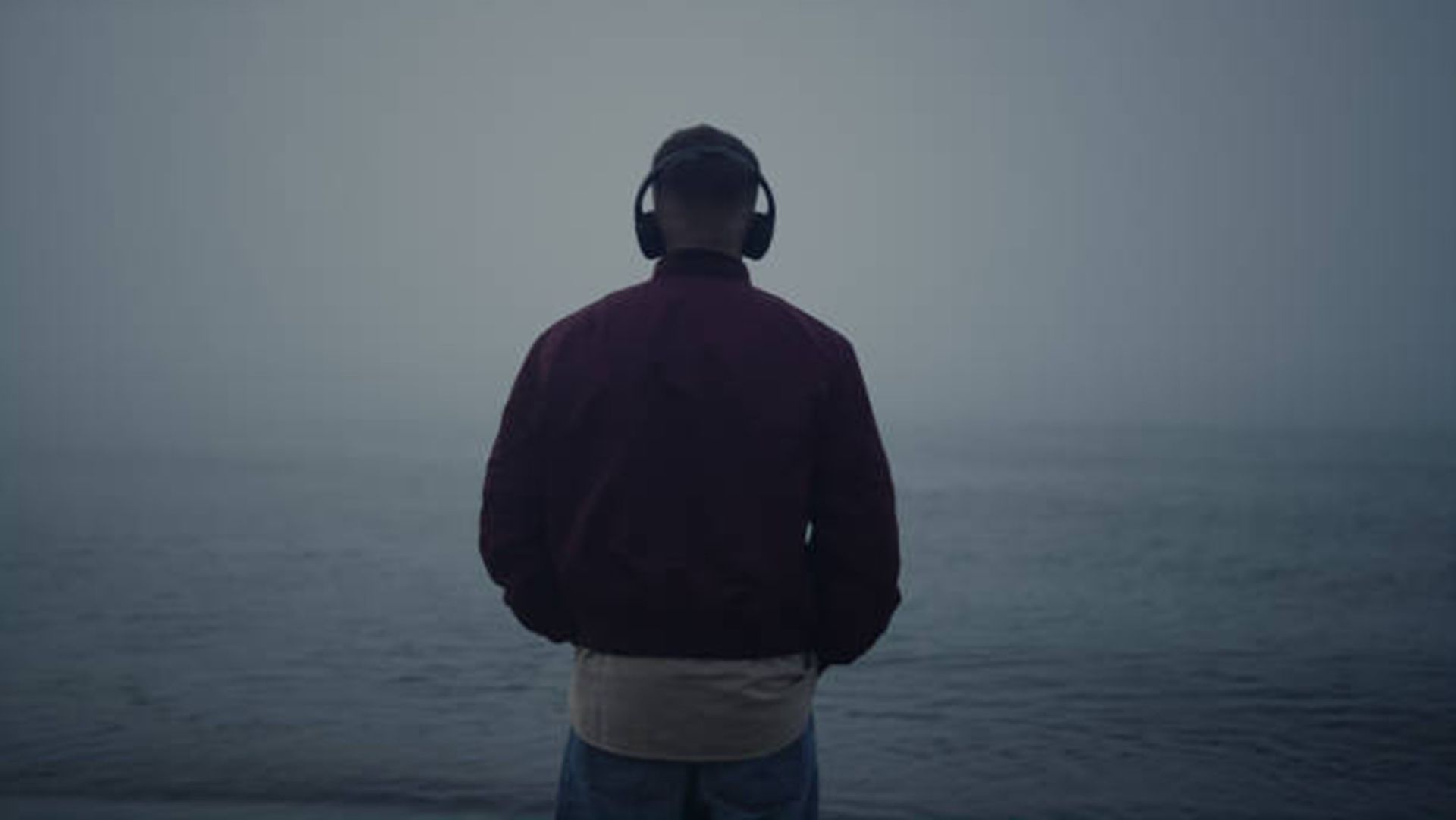 A man wearing headphones is standing on a beach looking at the ocean.