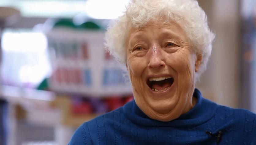 An elderly woman is laughing with her mouth open and wearing a blue sweater.
