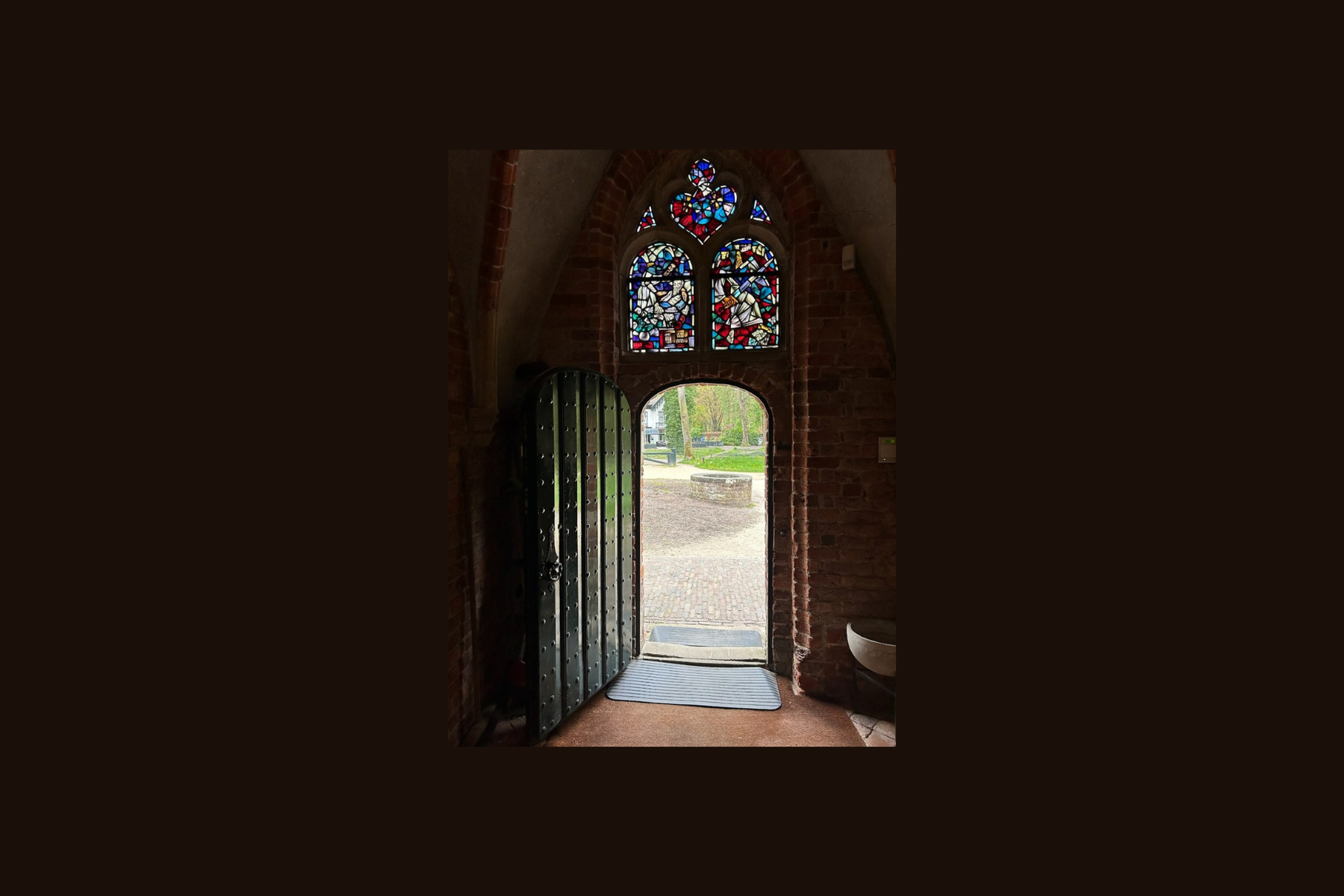 Open church door with stained-glass window above, letting in bright light and view of outside.