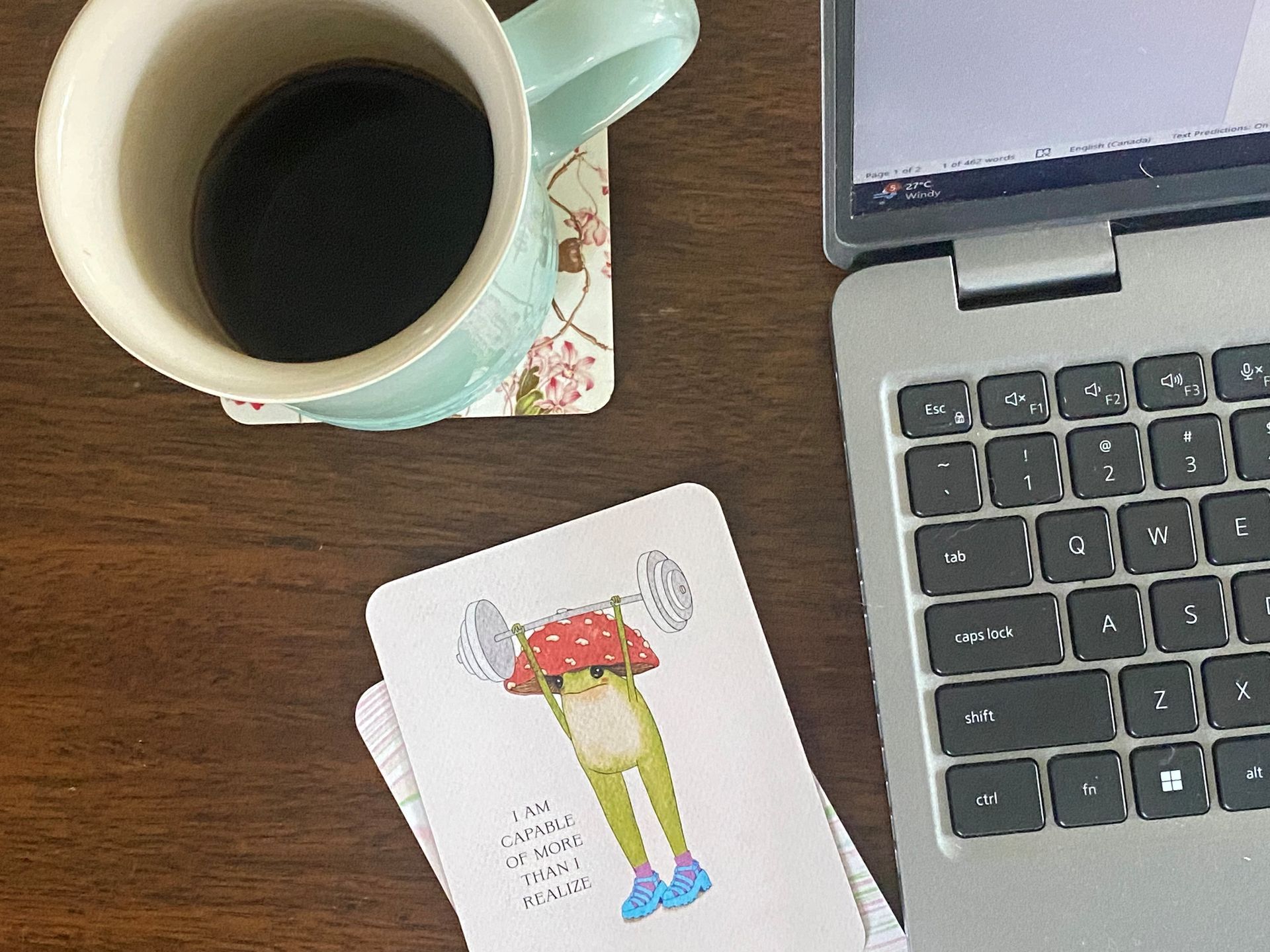 A cup of coffee next to a laptop and a card with a frog on it