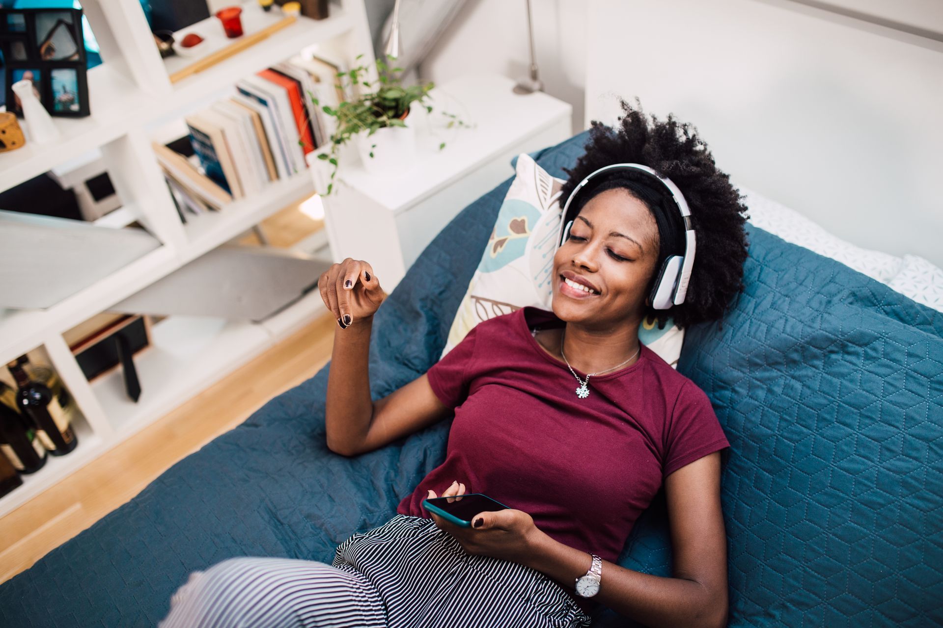A woman is laying on a bed wearing headphones and holding a cell phone.