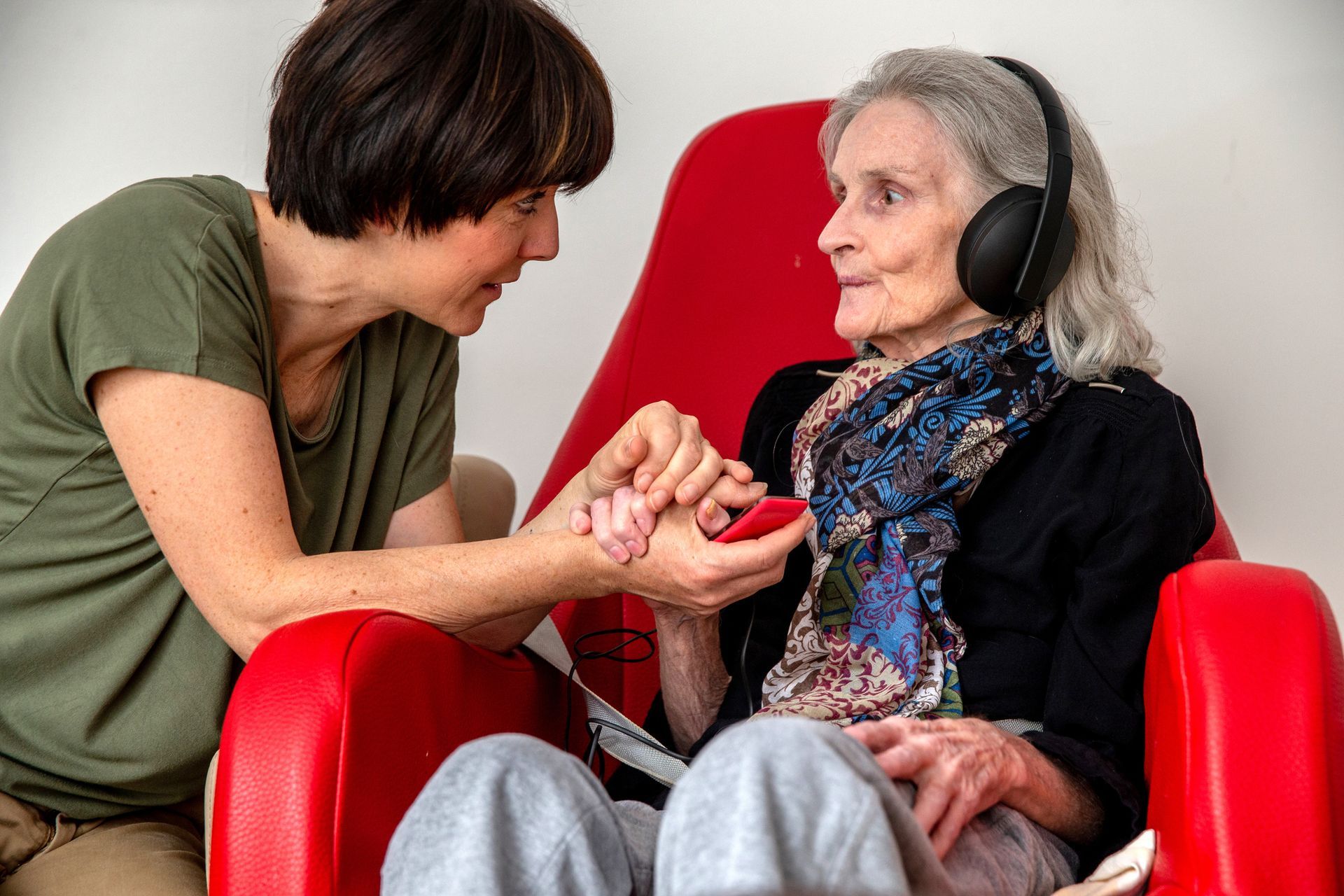 A woman is holding the hand of an elderly woman wearing headphones.