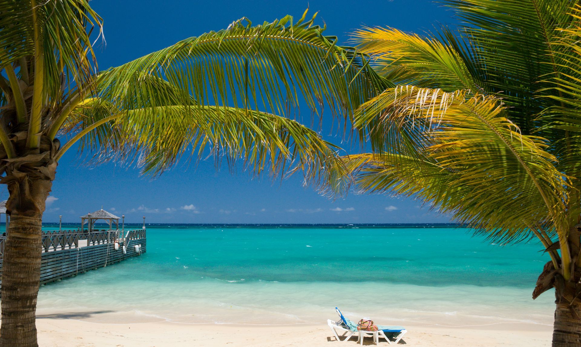 A beach with palm trees and chairs and a pier in the background.