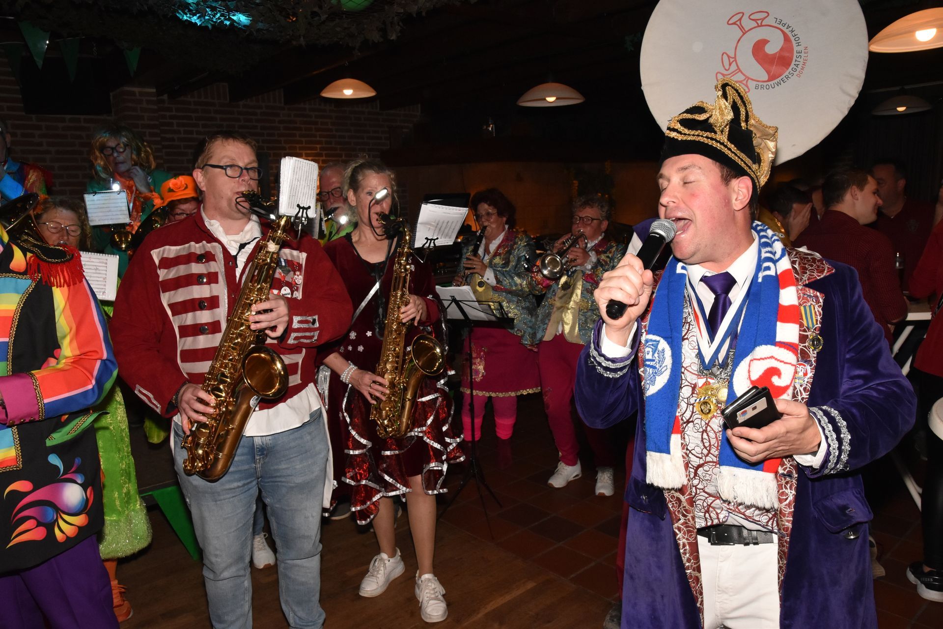 Binnen zitten twee mensen in carnavalskostuum, met boeken en een sjaal in de hand. Ze dragen hoeden met veren.