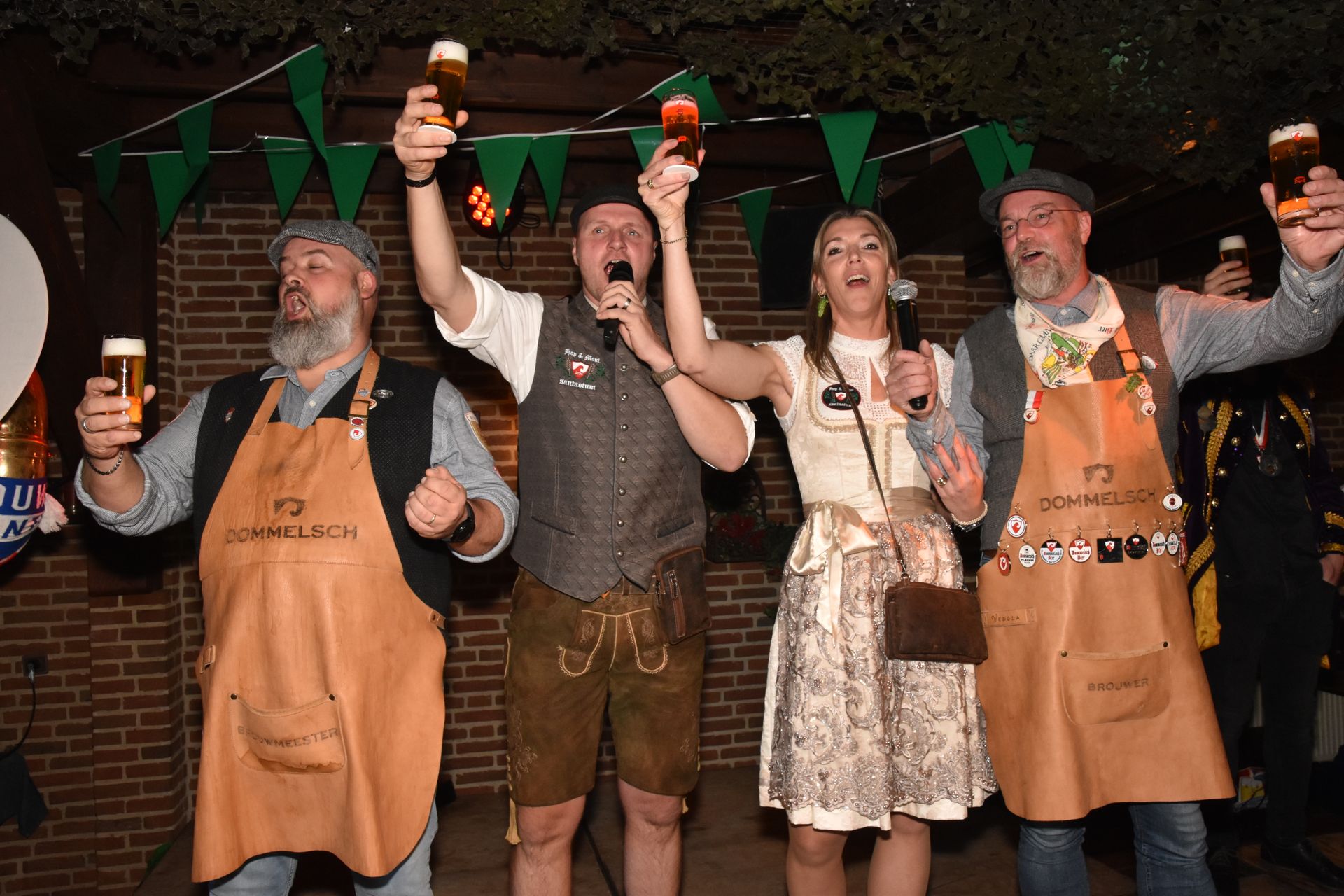 Mensen vieren feest op het podium, met vlaggen en bierpullen. Een biervat staat vooraan. Feestelijk evenement met lichtjes.