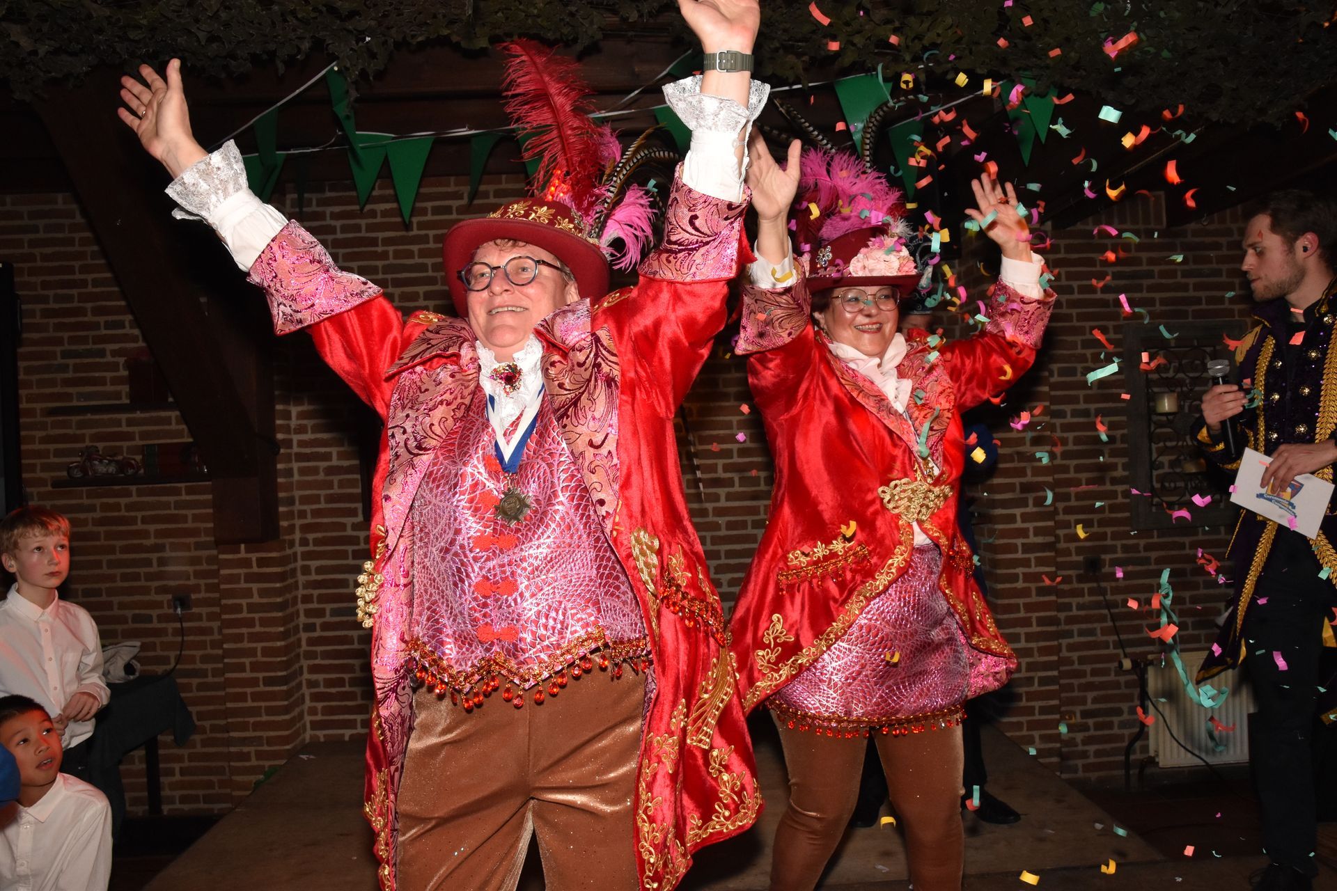 Twee mensen in feestelijke outfits, lachend bij een raam. De een in het groen, de ander in het blauw, met bijpassende hoeden.