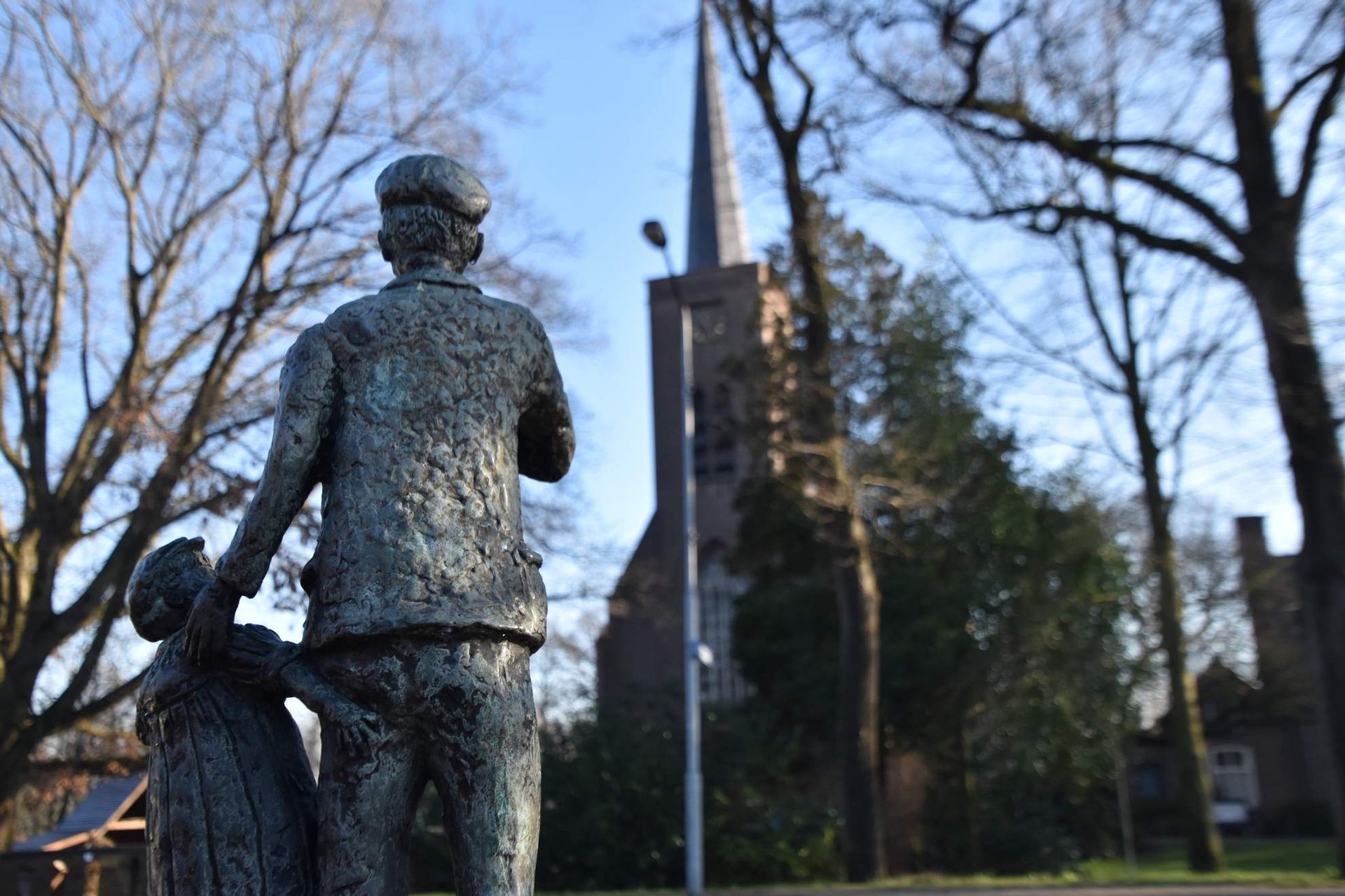 Bronzen beeld van een persoon die de hand van een kind vasthoudt, met de kerktoren op de achtergrond.