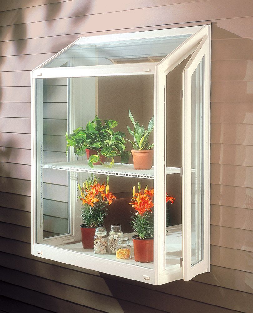 White, glass-paned wall greenhouse with two shelves holding potted plants, attached to a light brown wall. One door is open.