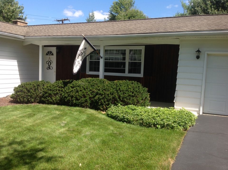 A one-story house with white siding and a brown roof. A manicured lawn features green bushes and a flag.