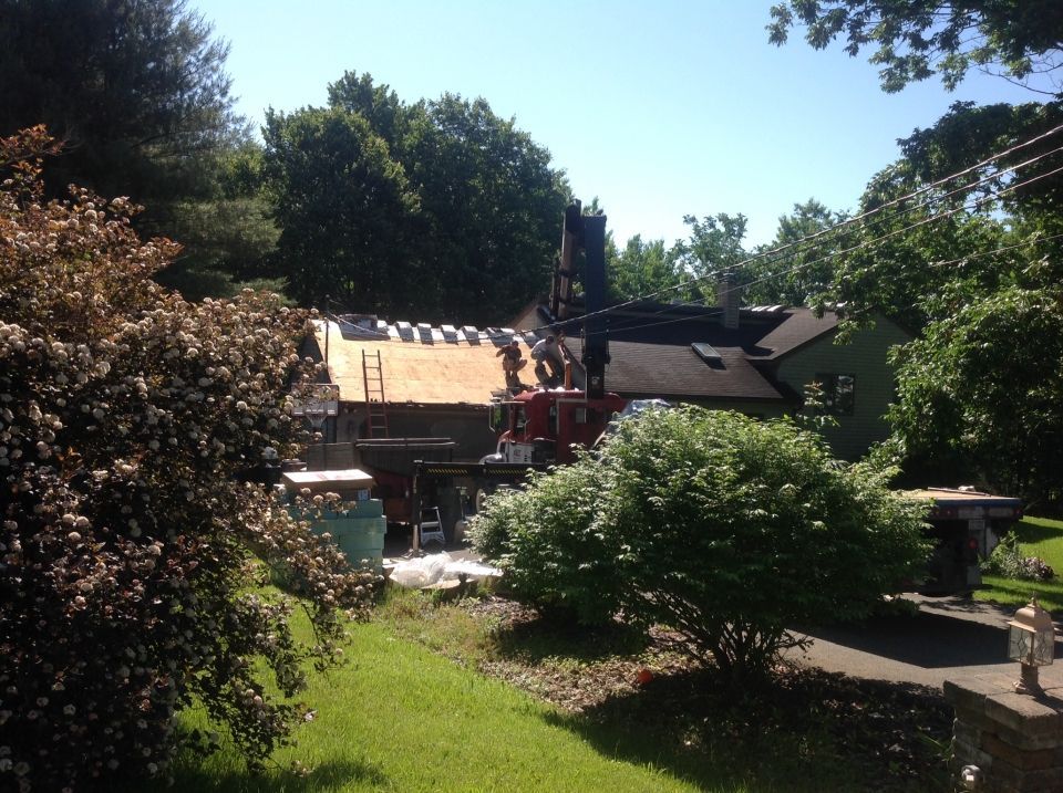 Roofers working on a house, visible from a grassy lawn with surrounding trees. Partially completed roof is visible.