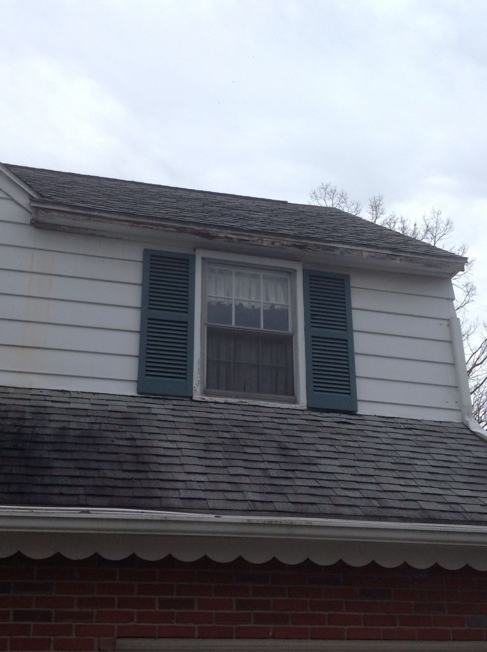 A white house with a dark gray roof and a dormer featuring a window with teal shutters. The sky is overcast.