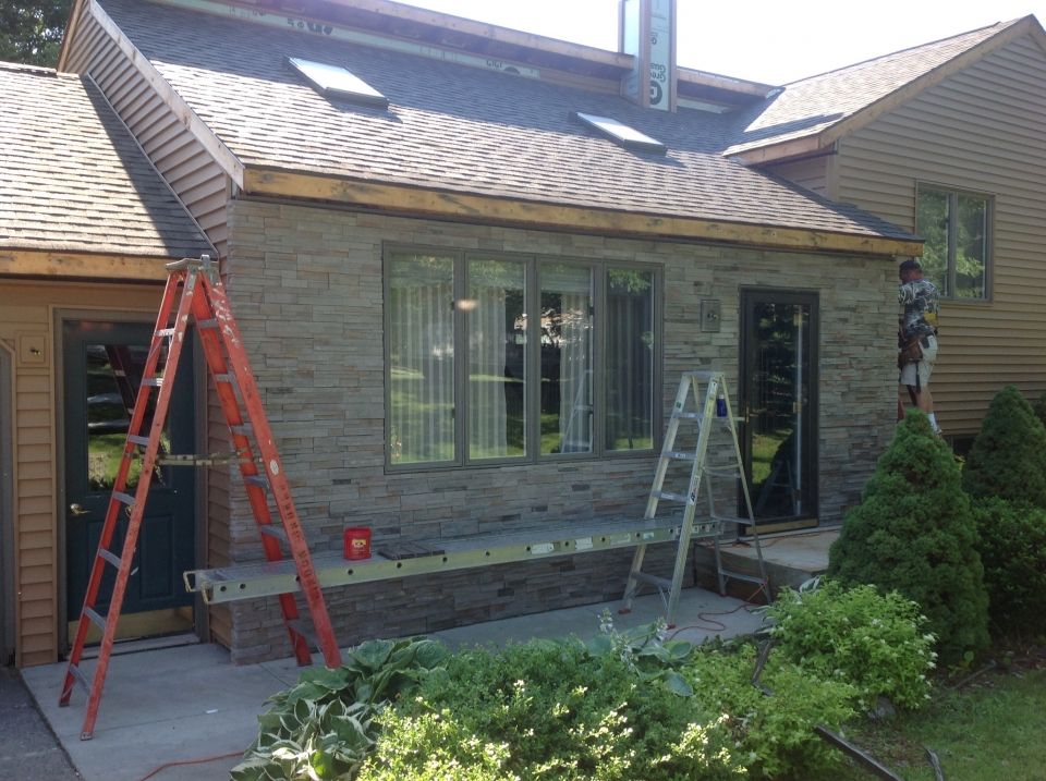 Exterior of a house undergoing construction. Gray stone siding is installed, ladders are propped against the wall, and a worker is on a ladder.