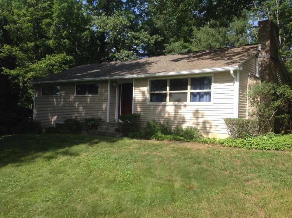 Ranch-style house with light siding, brown roof, and a brick chimney, set in a yard with green grass and trees.
