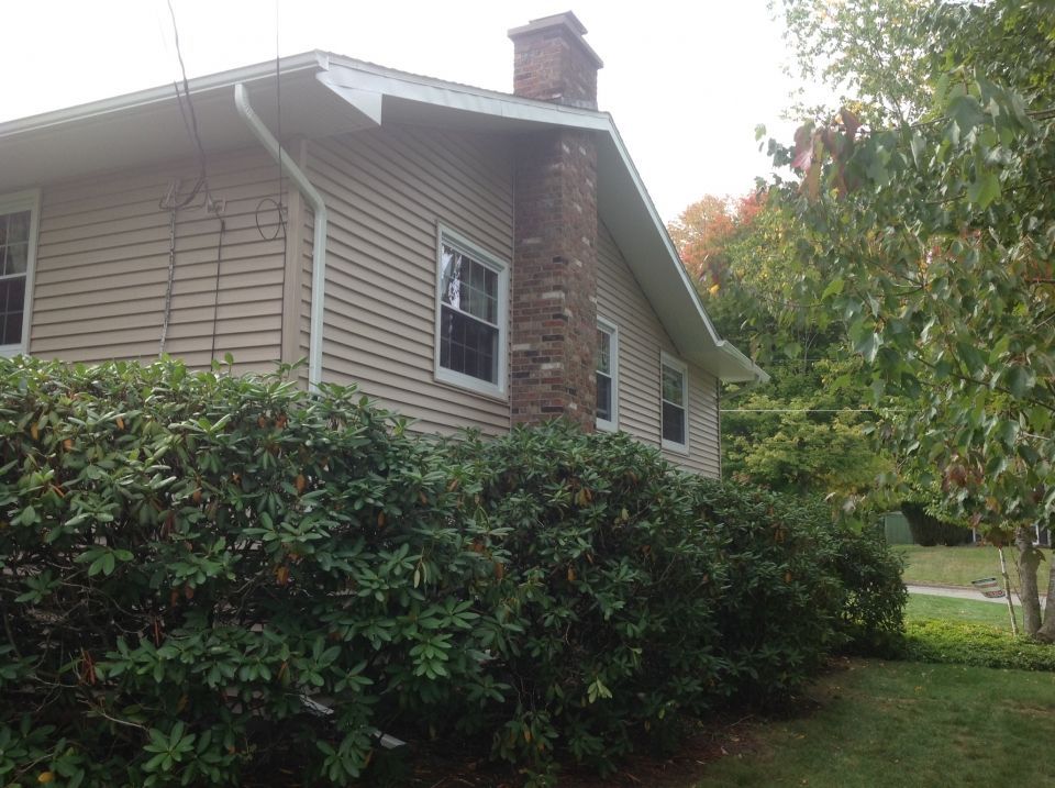 Beige-sided house with a brick chimney, green shrubs in front. Sunny outdoor setting.
