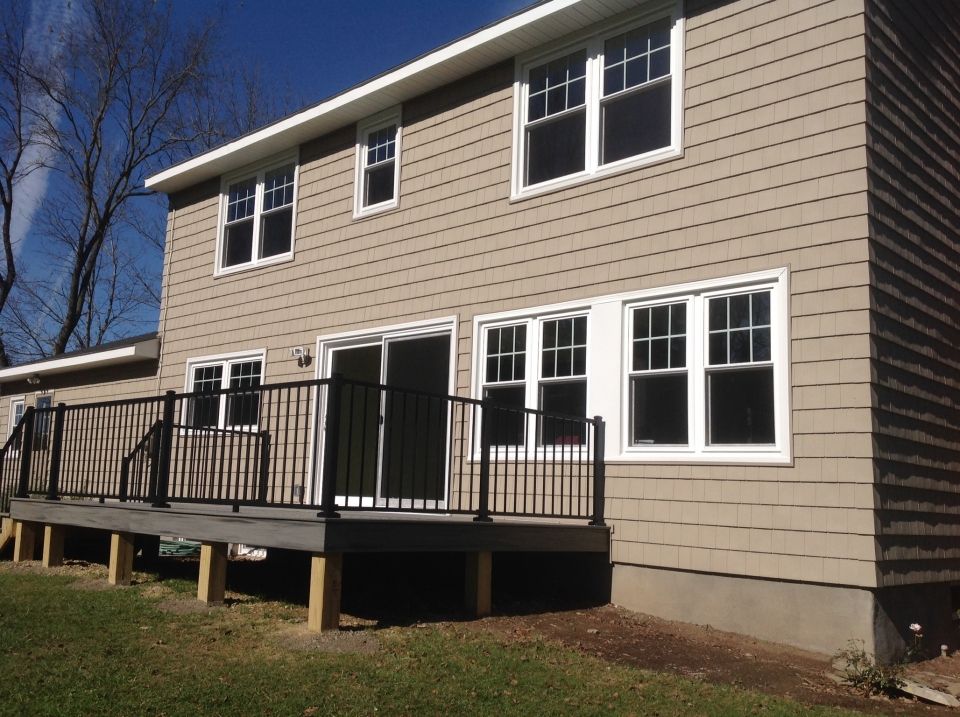 Two-story beige house with white-framed windows and a black-railed deck. A sunny, grassy yard is in front of the house.