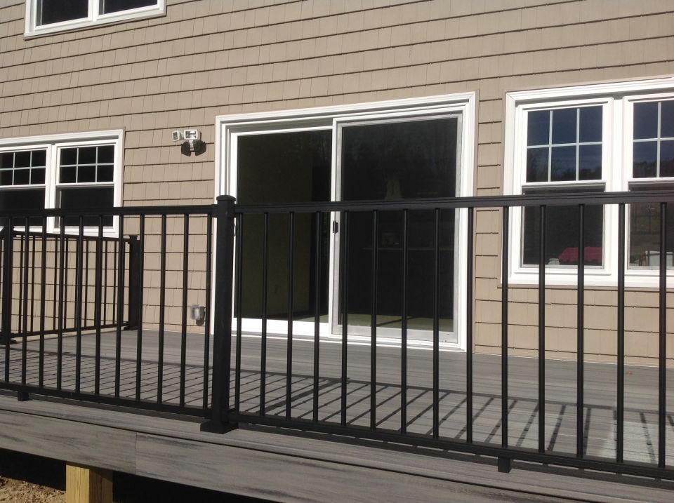 A black metal railing surrounds a gray deck in front of a home with beige siding, white windows, and a sliding glass door.