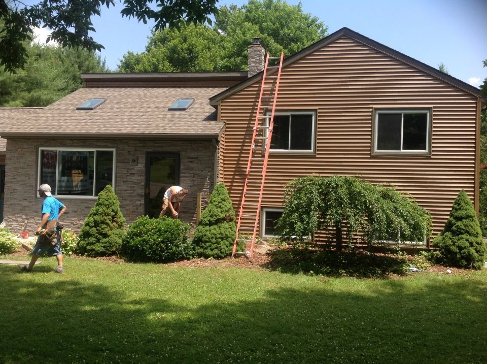 Workers on a house painting project: one walking, two near the bushes. Ladder against brown siding, stone facade, and manicured yard.