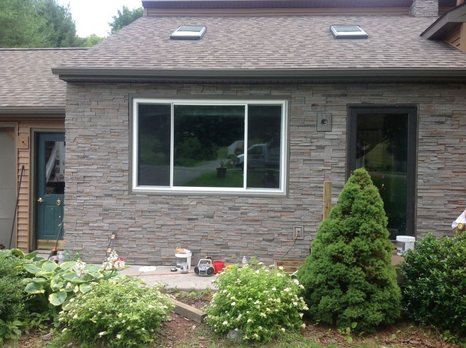 Exterior view of a house with stone veneer siding, featuring a large sliding window and a green front door. Bushes and small trees are in front.