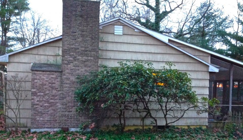 Tan siding house with brick chimney, surrounded by leafless trees and shrubs.