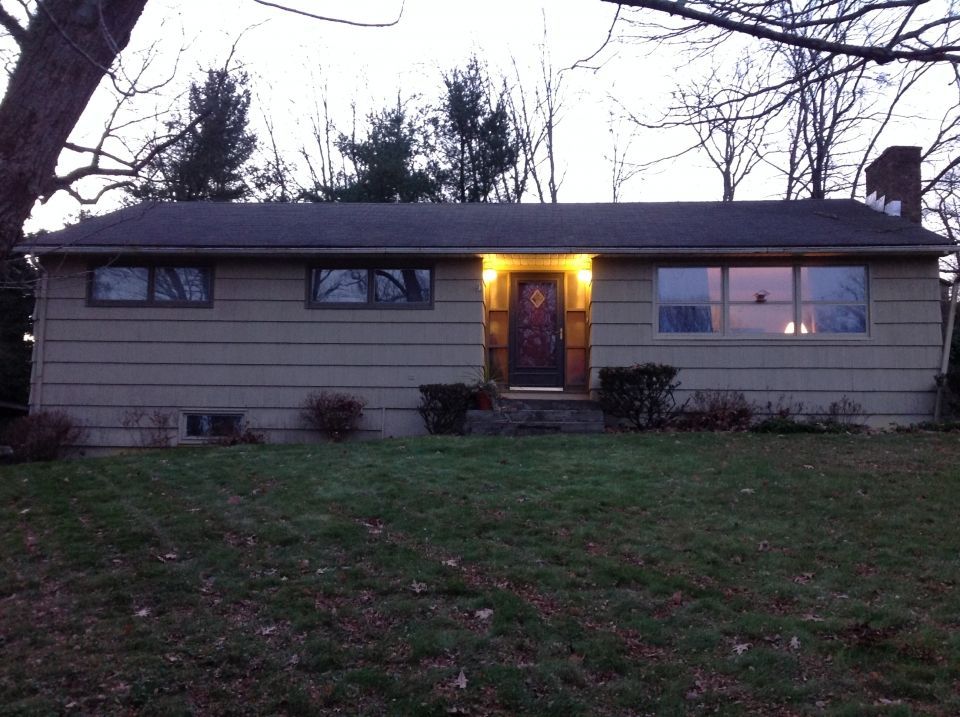 A single-story beige house with lit windows and a front door, set in a yard with brown grass.